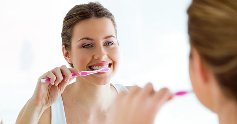 Woman looking in the mirror brushing her teeth and practicing dental quarantine tips.